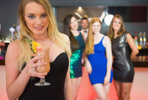 Blonde attractive woman holding cocktail standing in front of her friends and looking at camera