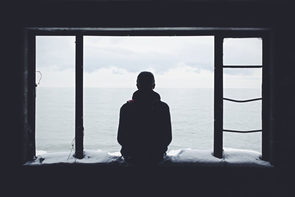 A lonely man sitting on a snow covered windowsill looking out over water